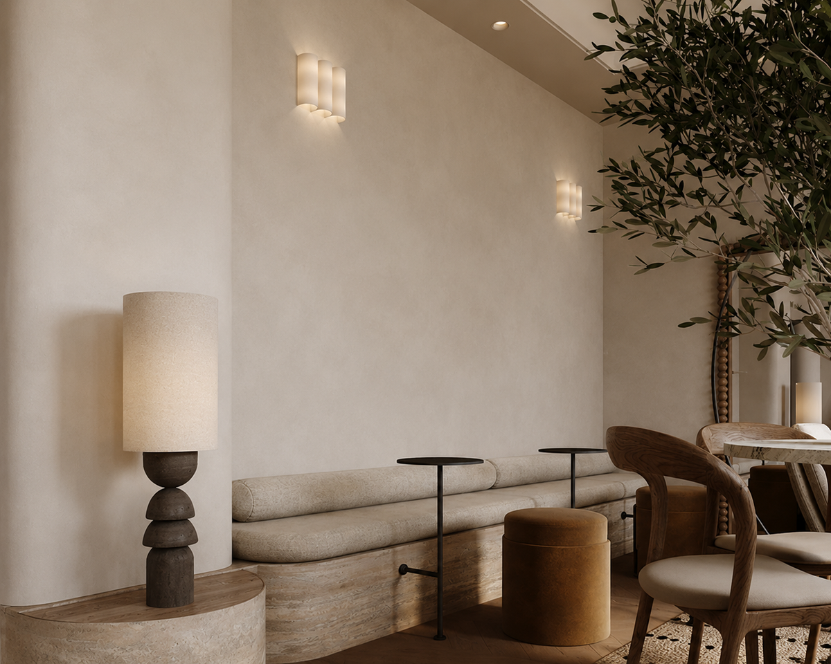 A modern interior scene with a beige upholstered bench against a textured cream wall, a sculptural table lamp on a round side table, small black side tables, a brown cylindrical ottoman, wooden dining chairs, and a leafy tree in the background.